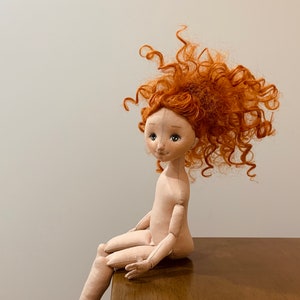 May include: A handmade doll with a beige fabric body and a curly, bright orange wig. The doll is sitting on a wooden surface with its legs dangling off the edge.