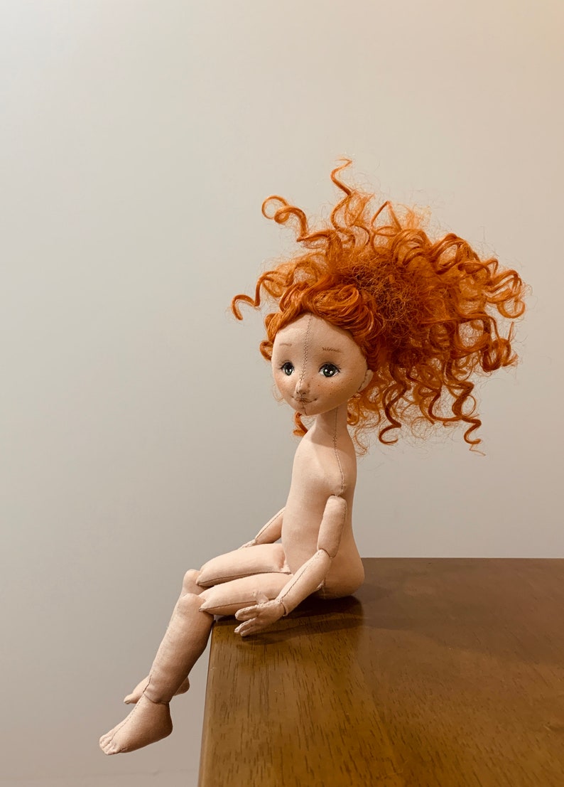 May include: A handmade doll with a beige fabric body and a curly, bright orange wig. The doll is sitting on a wooden surface with its legs dangling off the edge.