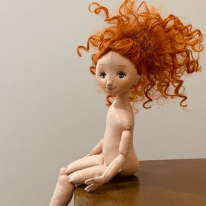 May include: A handmade doll with a red curly wig, sitting on a wooden surface. The doll has a stitched face with brown eyes and a stitched smile.