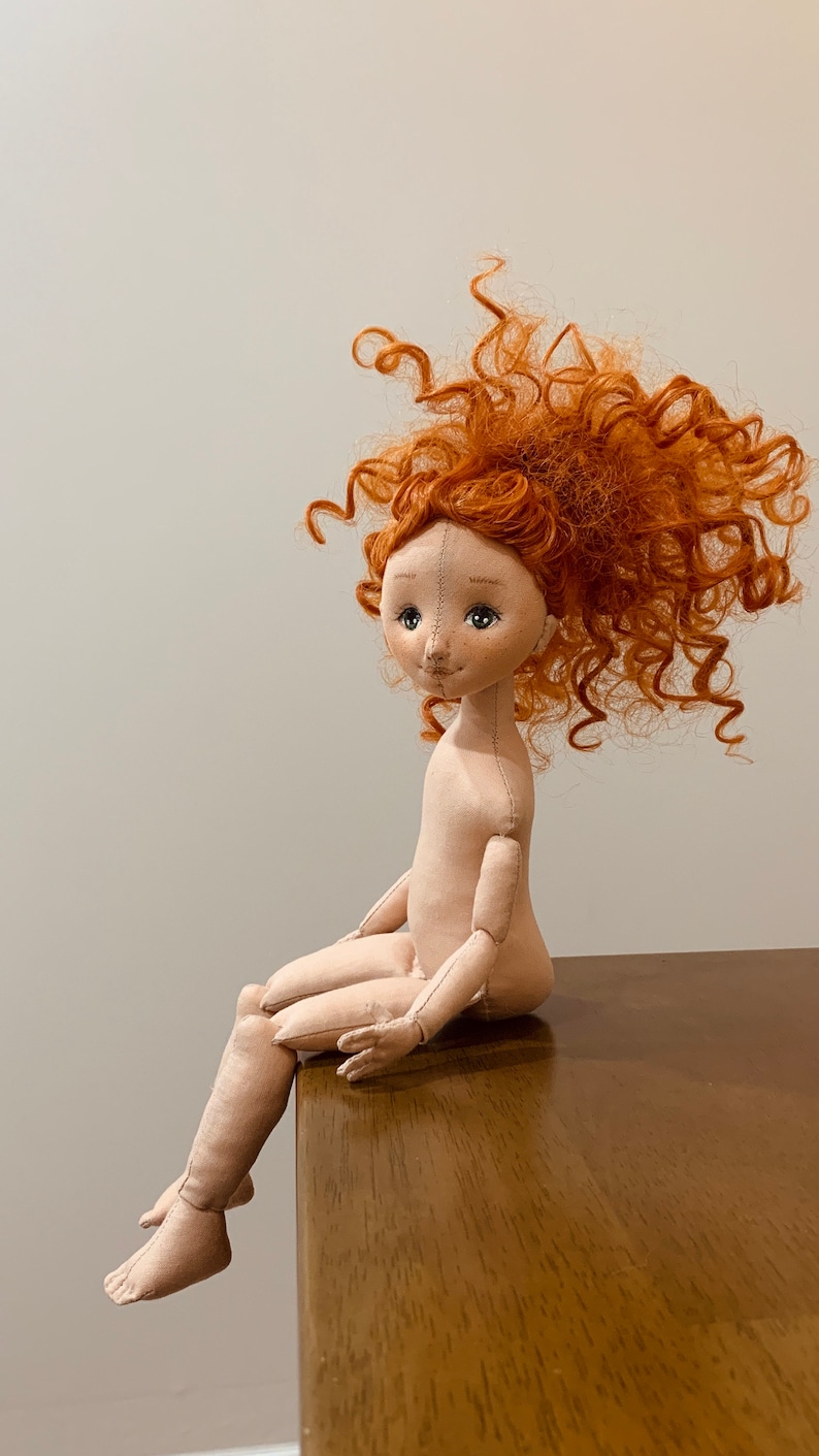 May include: A handmade doll with a red curly wig, sitting on a wooden surface. The doll has a stitched face with brown eyes and a stitched smile.