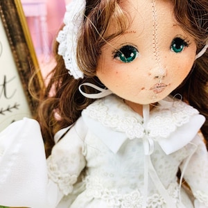 May include: A handmade doll with long brown hair, wearing a white dress with lace trim and a white bonnet. The doll has green eyes and a stitched smile.