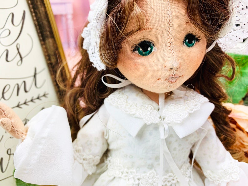 May include: A handmade doll with long brown hair, wearing a white dress with lace trim and a white bonnet. The doll has green eyes and a stitched smile.
