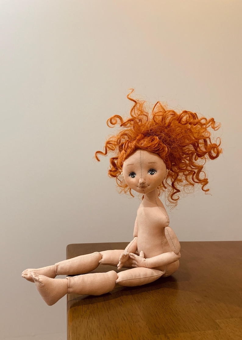 May include: A handmade doll with a red curly wig, sitting with its legs crossed. The doll has a light brown complexion and painted eyes.