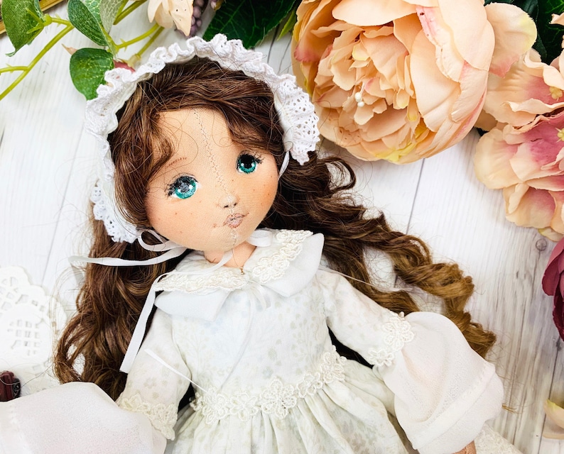 May include: A handmade doll with brown hair, wearing a white dress with lace trim and a white lace bonnet. The doll has blue eyes and a painted face.