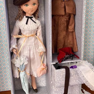 Könnte beinhalten: Eine Puppe mit braunem Barett, weißem Kleid und schwarzen Schuhen steht in einer Box. Die Puppe hält einen kleinen Sonnenschirm und eine rosa Handtasche. Ein zweites Fach enthält einen braunen Mantel und weiteres Zubehör.