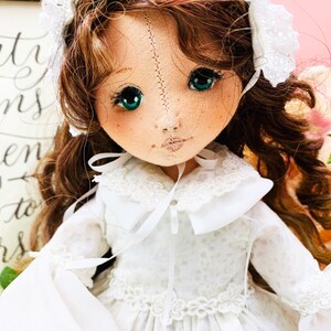 May include: A handmade doll with long brown curly hair, wearing a white dress with lace trim and a white bonnet. The doll has green eyes and a stitched face.