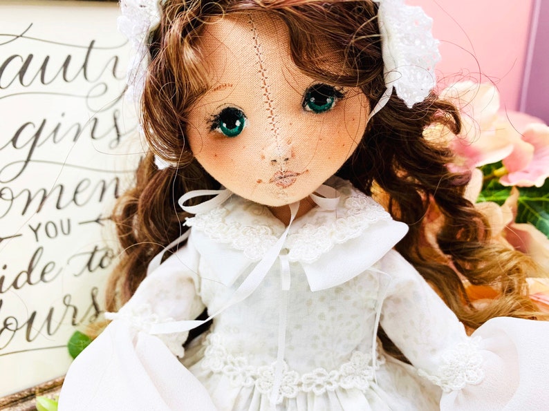 May include: A handmade doll with long brown curly hair, wearing a white dress with lace trim and a white bonnet. The doll has green eyes and a stitched face.