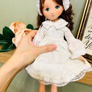 May include: A handmade doll with brown hair, a white dress with lace trim, and yellow shoes. The doll is wearing a white lace bonnet.