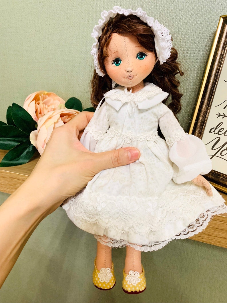 May include: A handmade doll with brown hair, a white dress with lace trim, and yellow shoes. The doll is wearing a white lace bonnet.