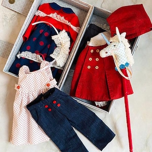May include: A selection of doll clothes and accessories presented in a grey box. The outfits include a red and navy blue dress with white lace, a pink polka dot dress, and navy blue trousers. A red cape and a unicorn stick horse are also included.