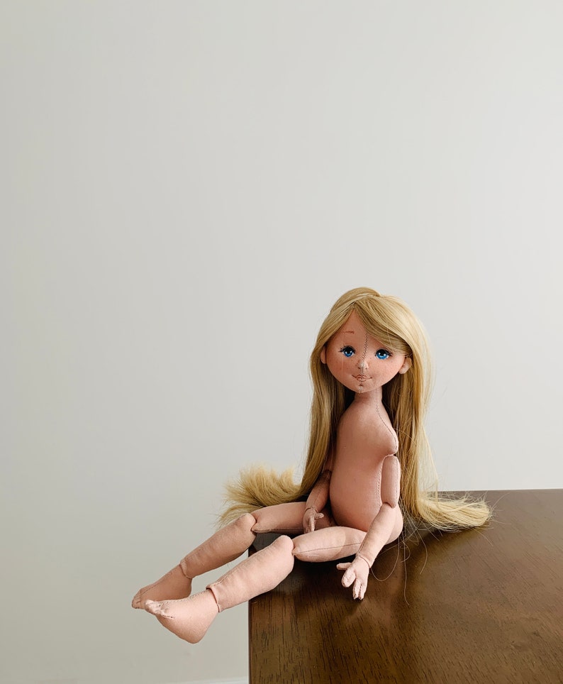May include: A handmade doll with long blonde hair, blue eyes, and a pink fabric body. The doll is sitting on a wooden surface.