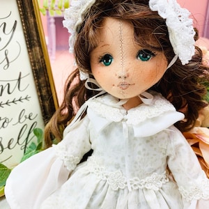 May include: A white doll with brown hair wearing a white dress with lace trim and a white bonnet. The doll has green eyes and a stitched face.