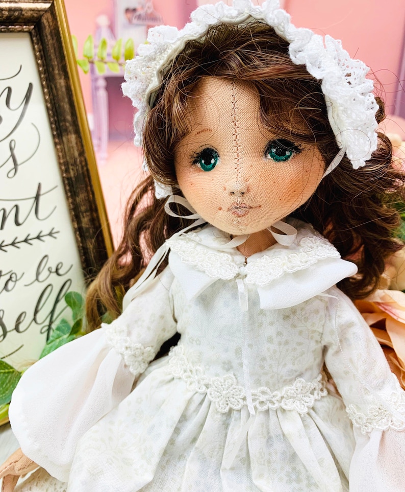 May include: A white doll with brown hair wearing a white dress with lace trim and a white bonnet. The doll has green eyes and a stitched face.