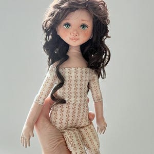 May include: A handcrafted doll with long, wavy dark brown hair, green eyes, and freckles. The doll wears a cream-coloured jumpsuit featuring a repeating vertical pattern of small brown floral sprigs, with three-quarter length sleeves and a straight neckline.
