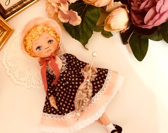 May include: A handmade doll with blonde hair, wearing a pink bonnet, a black and white polka dot dress with a pink lace trim, and black shoes. The doll is holding a lace parasol.