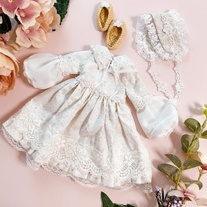 Puede incluir: Un vestido color crema con un sutil estampado floral, que presenta mangas largas con puños de volantes, un cuello adornado con encaje y un dobladillo ancho de encaje. El conjunto incluye un gorro a juego con encaje y un par de zapatos pequeños con lunares amarillos.