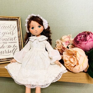 May include: A white porcelain doll with brown hair wearing a white dress with lace trim and a white bonnet. The doll is sitting on a wooden shelf with pink and white flowers behind it. The doll has blue eyes and a painted smile. The shelf has a framed sign with the text "Beauty begins the moment you decide to be yourself."