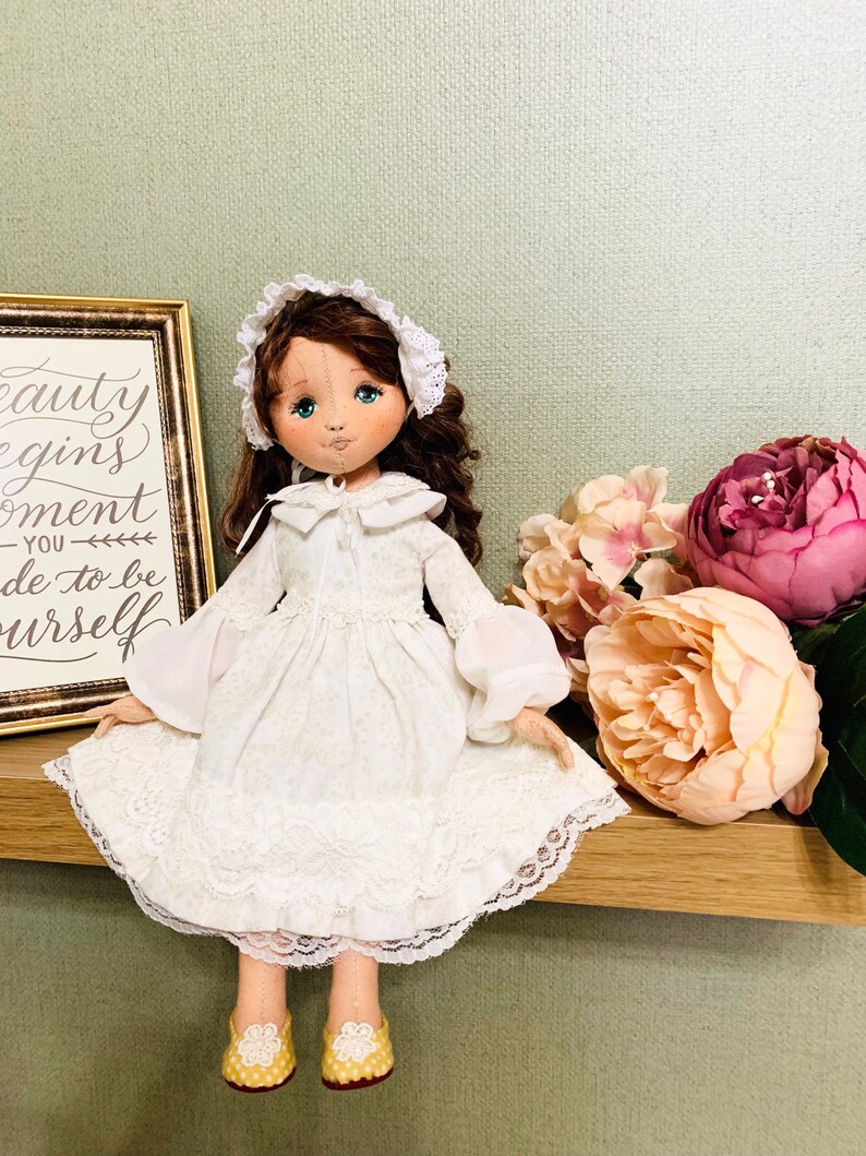 May include: A white porcelain doll with brown hair wearing a white dress with lace trim and a white bonnet. The doll is sitting on a wooden shelf with pink and white flowers behind it. The doll has blue eyes and a painted smile. The shelf has a framed sign with the text "Beauty begins the moment you decide to be yourself."