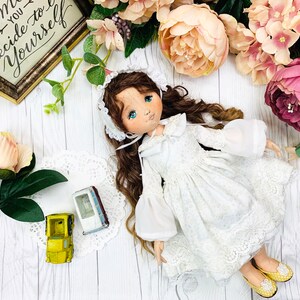 May include: A white fabric doll with brown hair wearing a white dress with lace trim and a white bonnet. The doll is lying on a white wooden surface with flowers and a framed sign that reads "Live in the moment, you decide to be yourself".