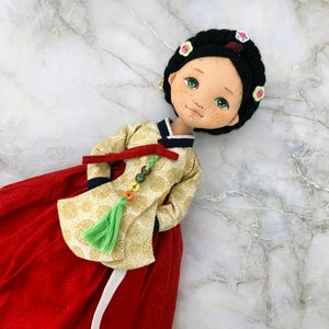 以下が含まれることがあります： 伝統的な韓国のハンボクを着た手作りの人形。人形は色白で、緑色の目とそばかすがあります。ハンボクは金色のスカートと赤いスカート、緑色のタッセルです。人形は花のアクセントが付いた黒い髪をしています。