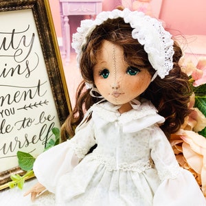 May include: A porcelain doll with brown hair wearing a white dress and a white lace bonnet. The doll has blue eyes and a painted smile. The doll is sitting on a floral background.