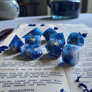May include: A collection of blue and gold resin dice, including a pyramid and various multi-sided dice, are arranged on a vintage paper with text. The dice have gold accents and are scattered with small blue decorative elements.