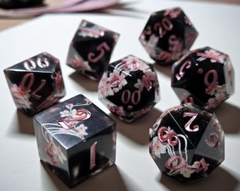 May include: Set of seven black polyhedral dice with pink floral designs and numbers. The dice are for role-playing games and have a glossy finish. The numbers are in a pink font.
