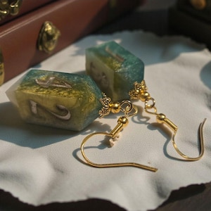 May include: A pair of gold-toned earrings with a unique design. Each earring features a rectangular, translucent charm with a teal and gold color scheme, and the number "2" visible. The earrings are displayed on a white cloth.