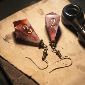 May include: A pair of dangle earrings featuring elongated, diamond-shaped resin charms in a marbled red and white pattern. Each charm displays a gold-colored numeral, a three and a two, attached to antique brass-toned metal hooks with small beads.