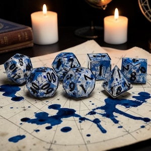 May include: A set of seven polyhedral dice featuring a white base with blue ink-like splatters and black numbers. The dice set includes a d20, d12, d10, d8, d6, d4, and a percentile die, all arranged on a piece of parchment paper with blue ink stains.