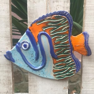 Può includere: Un ornamento di pesce in ceramica dipinto di blu, arancione e verde. Il pesce è appeso a una recinzione di legno bianco.