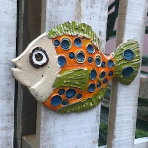 Könnte beinhalten: Keramische Fisch-Wanddekoration mit orangefarbenen, blauen und grünen Akzenten. Der Fisch hat ein cremefarbenes Gesicht und ein strukturiertes Design. Er wird an einer Schnur aus Naturfaser aufgehängt und hängt an einem weißen Holzzaun.
