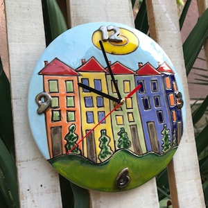May include: A colorful ceramic clock with a design of houses and a sun. The clock face has Roman numerals and the hands are black and red.