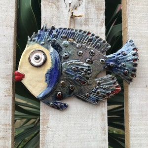 Puede incluir: Un adorno de pared de cerámica con forma de pez pintado en azul, blanco y rojo. El pez tiene un ojo grande y redondo y una boca pequeña y roja.