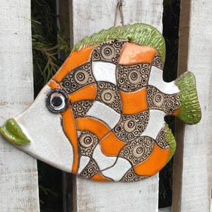 Op de afbeelding: Een keramische vis wanddecoratie met een wit, oranje en bruin geometrisch patroon. De vis heeft een groene vin en een zwart oog.