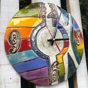 Puede incluir: Un colorido reloj de pared de cerámica con una esfera redonda. El reloj presenta un diseño segmentado con secciones amarillas, naranjas, rojas, azules y moradas. Se ven las manecillas negras y un segundero rojo. Se incluyen patrones decorativos.