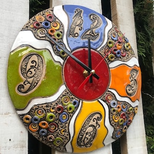May include: A colourful ceramic clock with Roman numeral markings. The clock face is divided into six sections, each painted in a different colour: red, orange, yellow, green, blue, and white. The clock hands are black and red.