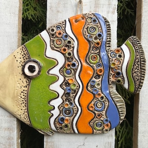 May include: A ceramic fish wall hanging with a colorful abstract design. The fish is painted in shades of green, orange, blue, white, and black. It has a large eye and a textured surface.