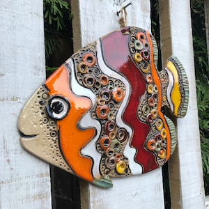 Puede incluir: Un adorno de pared de cerámica con forma de pez con un diseño colorido. El pez está pintado en tonos de naranja, rojo, amarillo, blanco y marrón. Tiene una superficie texturizada con pequeños círculos y remolinos.