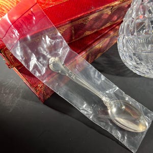 Puede incluir: Una cuchara de plata en una bolsa de plástico transparente, con detalles ornamentados en el mango y el cuenco. La cuchara se encuentra sobre una caja roja con detalles dorados y un cuenco de cristal tallado.