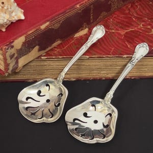 Puede incluir: Dos cucharas de servir antiguas de plata con mangos ornamentados y cuencos calados. Las cucharas descansan sobre una superficie oscura con un libro vintage en el fondo. Las cucharas tienen un diseño floral en el cuenco.