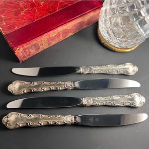 May include: Four antique silver knives with ornate handles and shiny blades are arranged on a dark surface. The handles feature detailed scrollwork and a decorative end. A red book and a crystal glass are in the background.