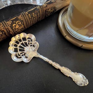 May include: A silver-colored, ornate serving spoon with a fan-shaped head and a detailed handle. The spoon is likely vintage, with intricate filigree patterns. The spoon is on a dark surface, with a book and candle in the background.