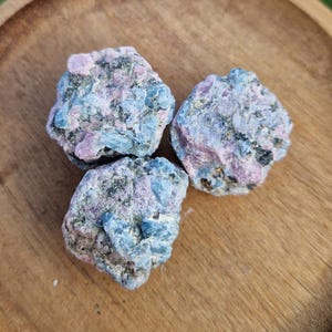 May include: Three rough, raw, blue kyanite crystals with pink inclusions. The crystals are clustered together on a wooden surface.