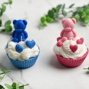 May include: Two cupcake candles, one blue and one pink, each topped with a teddy bear figurine and hearts. The blue cupcake has a blue bear and blue hearts, while the pink cupcake has a pink bear and pink hearts.