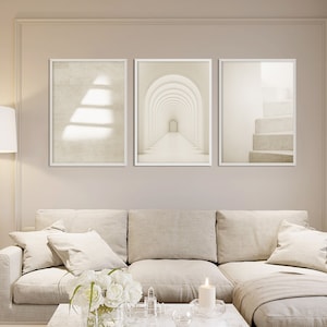 May include: A set of three vertical wall art prints in white frames. The left print shows a textured beige surface with light shadows. The centre print depicts a series of repeating white arches. The right print features a staircase in neutral tones.