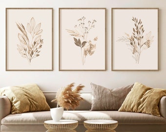 May include: Three framed prints with a minimalist design featuring brown line drawings of botanicals on a cream background. The prints are hung on a white wall above a light brown couch with throw pillows and a vase with dried pampas grass.