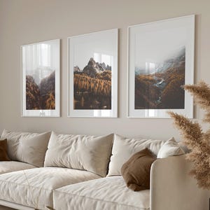 May include: Three vertical framed wall art prints featuring mountain landscapes with autumn forests and rocky peaks, displayed in white frames above a beige sofa with brown accent pillows and a round wooden coffee table with books.