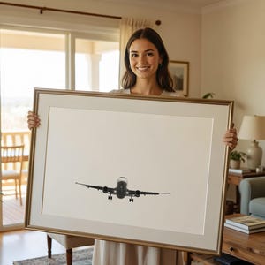 May include: A framed print showing a black and white aeroplane in flight. The artwork is presented in a gold-coloured frame. The image is held by a person wearing a light-coloured dress.