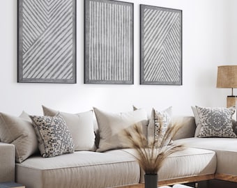 May include: Three framed abstract art prints with gray and white geometric designs hang above a beige sectional sofa with decorative pillows. A wooden coffee table holds a vase of dried plants and an open book.